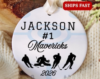 May include: A round white ceramic ornament featuring a light blue hockey rink graphic, the name JACKSON, the number #1, the team name Mavericks, and the year 2026. Four black silhouettes of hockey players are printed at the bottom.