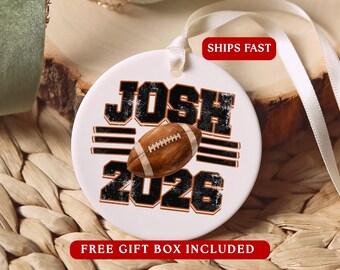 May include: A round white ceramic ornament featuring the name JOSH in black distressed lettering above a brown football graphic and the year 2026 in black distressed lettering below. The ornament includes a white ribbon for hanging.