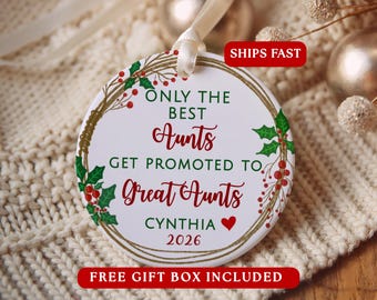 May include: A round white ceramic ornament featuring a gold wreath design with green holly leaves and red berries. The text reads: ONLY THE BEST Aunts GET PROMOTED TO Great Aunts CYNTHIA with a red heart and the year 2026 in red ink.