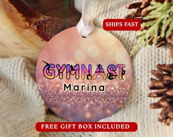May include: A round, flat ornament featuring a gradient background of soft pink and orange with a bokeh effect. The word GYMNAST is printed in a bold, multi-coloured font with black silhouettes of gymnasts, followed by the name Marina in black.