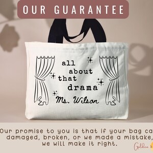 May include: A white canvas tote bag with black handles featuring a black graphic print of stage curtains on both sides. The center text reads all about that drama and Ms. Wilson, accented with small star symbols. The bag has a rectangular shape.