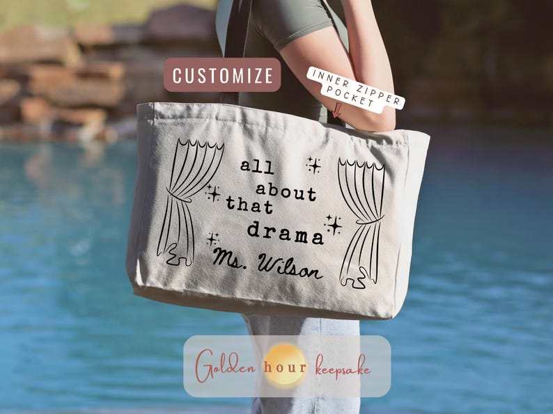 May include: A beige tote bag with black text that reads "all about that drama Ms. Wilson" and curtain graphics. The bag has an inner zipper pocket and is being held outdoors.