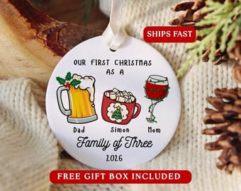 May include: A round white ceramic Christmas ornament with a ribbon hanger. It features illustrations of a beer mug, a hot cocoa mug, and a wine glass. Text reads: OUR FIRST CHRISTMAS AS A FAMILY OF THREE, Dad, Simon, Mom, and 2026.