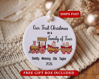 May include: A round white ceramic ornament featuring a red and gold train graphic with gingerbread characters. The text reads Our First Christmas as a Family of Four, Daddy, Mommy, Ella, Taylor, and 2026. It hangs from a cream-coloured ribbon.