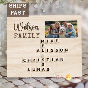May include: A wooden family photo frame with the name "Wilson Family" engraved at the top. A family photo is displayed in the frame, and names are spelled out with scrabble tiles. The frame includes a paw print.