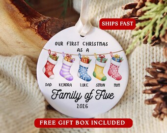 May include: A round white ceramic Christmas ornament hanging from a cream ribbon. It features five colourful stockings hanging on a string with the text: OUR FIRST CHRISTMAS AS A, DAD, KENDRA, LUKE, SIMON, MOM, Family of Five, and 2026.