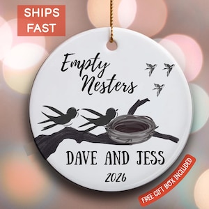 May include: A white ceramic ornament with a black and white illustration of birds, a nest, and the words "Empty Nesters," "Dave and Jess," and the year 2026. A gold string is attached for hanging.