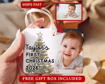May include: A rectangular white ornament featuring a watercolour Christmas tree with a star topper, the text Taylor's FIRST CHRISTMAS 2026, and a photograph of a smiling baby. The ornament includes a cream-coloured ribbon for hanging.
