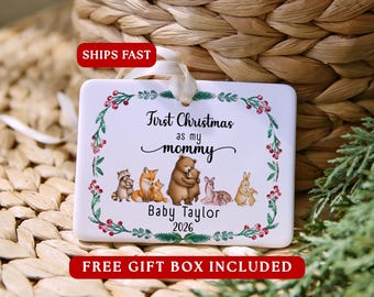 May include: A white rectangular ceramic ornament with a cream ribbon, featuring a watercolour illustration of forest animals and the text First Christmas as my mommy Baby Taylor 2026, framed by a green and red holly berry wreath.