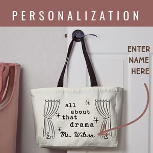 May include: Cream-colored tote bag with black handles, hanging on a white door. The bag features the text "all about that drama" with a name below, and illustrations of theater curtains. The word "PERSONALIZATION" is at the top.