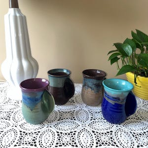 May include: Four ceramic mugs with integrated side handles, each featuring a unique multi-colored glaze. The mugs display various color combinations including purple and green, dark brown and blue, tan and blue, and a vibrant blue and turquoise.