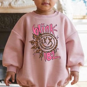 Puede incluir: Un bebé con un body rosa con el texto "Blink 182" en un diseño gráfico. El conjunto se combina con una diadema a juego, calcetines rosas y botas negras con cordones rosas. El bebé está de pie.