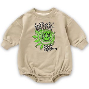 Puede incluir: Body de bebé color beige con mangas largas y cuello redondo. La parte delantera presenta un gráfico de carita sonriente verde neón con las palabras "blink" y "182" en un patrón de cuadros blanco y negro.