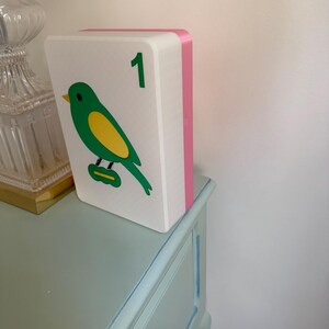 May include: A decorative white block with a green and yellow bird illustration and the number 1 in green. The block has a pink side and is resting on a light blue dresser.
