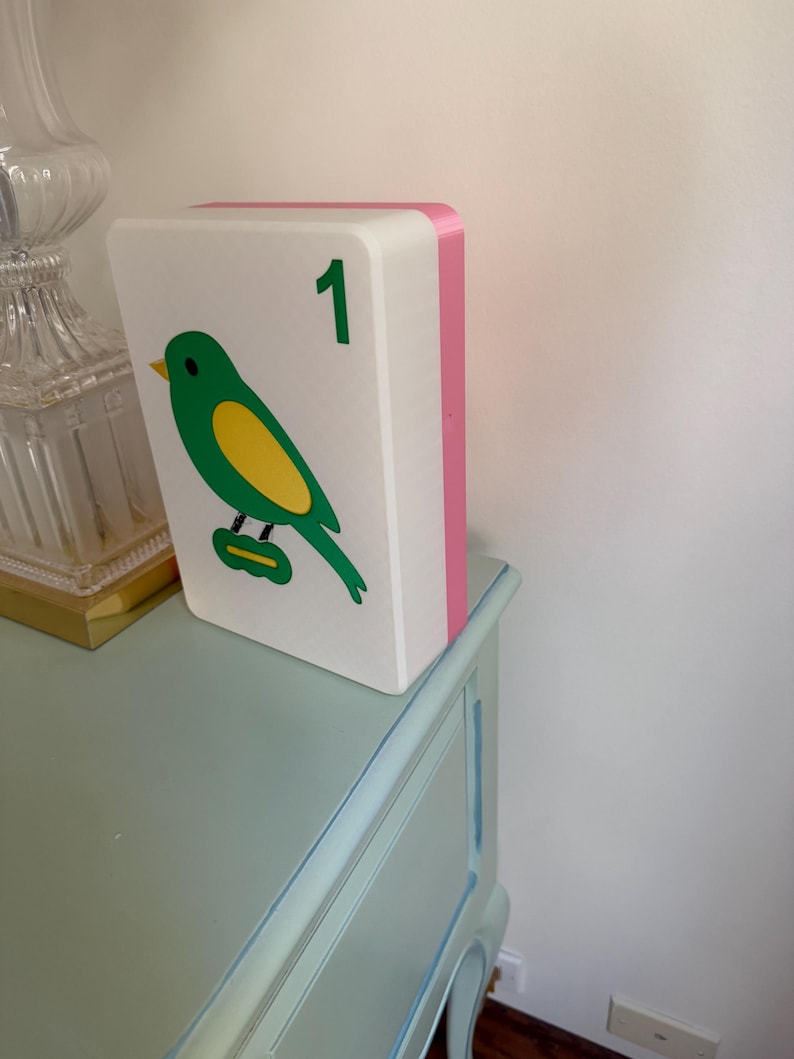 May include: A decorative white block with a green and yellow bird illustration and the number 1 in green. The block has a pink side and is resting on a light blue dresser.