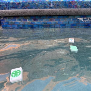 May include: Three small, white game cards with colorful designs floating in a pool of water. The cards have green, blue, and white accents. The pool has a blue tile border.