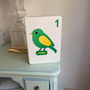 May include: A white, rectangular decorative block featuring a green bird with a yellow belly and a green number 1. The bird is perched on a green base. The block is displayed on a light blue table.