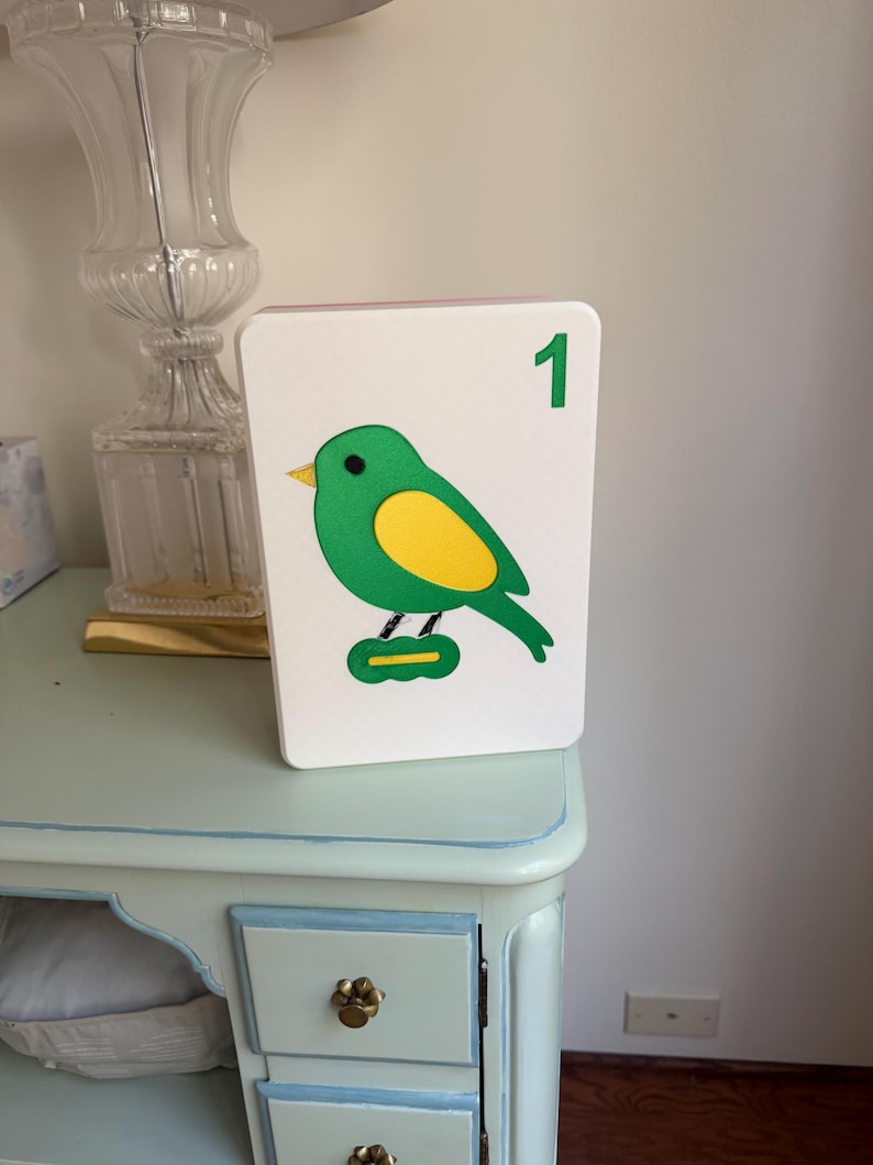 May include: A white, rectangular decorative block featuring a green bird with a yellow belly and a green number 1. The bird is perched on a green base. The block is displayed on a light blue table.