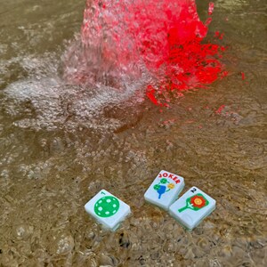 May include: Three white game tiles with colorful illustrations and the word "JOKER" floating in water. The tiles feature a green polka dot, a blue and yellow design, and a red and orange design. Red water is splashing in the background.