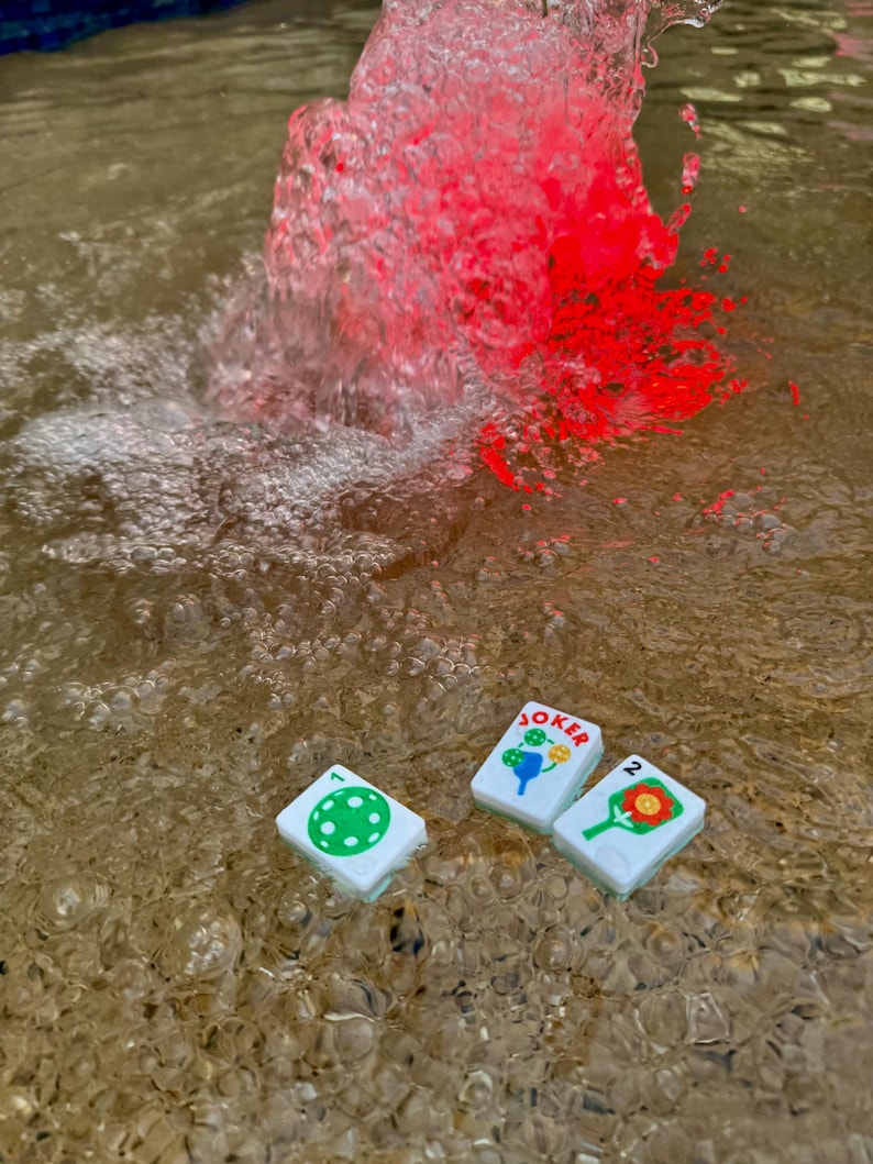 May include: Three white game tiles with colorful illustrations and the word "JOKER" floating in water. The tiles feature a green polka dot, a blue and yellow design, and a red and orange design. Red water is splashing in the background.