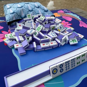 May include: A collection of Mahjong tiles with purple backs and white faces, scattered on a blue mat with a koi fish design. A matching blue pouch and a tile rack are also present. The tiles display various symbols and numbers.