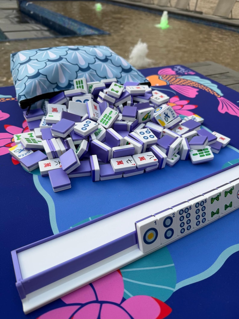 May include: A collection of Mahjong tiles with purple backs and white faces, scattered on a blue mat with a koi fish design. A matching blue pouch and a tile rack are also present. The tiles display various symbols and numbers.