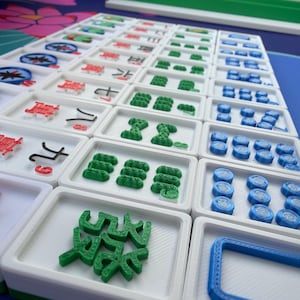 May include: A set of Mahjong tiles arranged on a blue surface. The tiles are white with green, red, and blue accents. The game pieces feature various symbols and shapes, including Chinese characters and geometric designs. The set is ready for play.