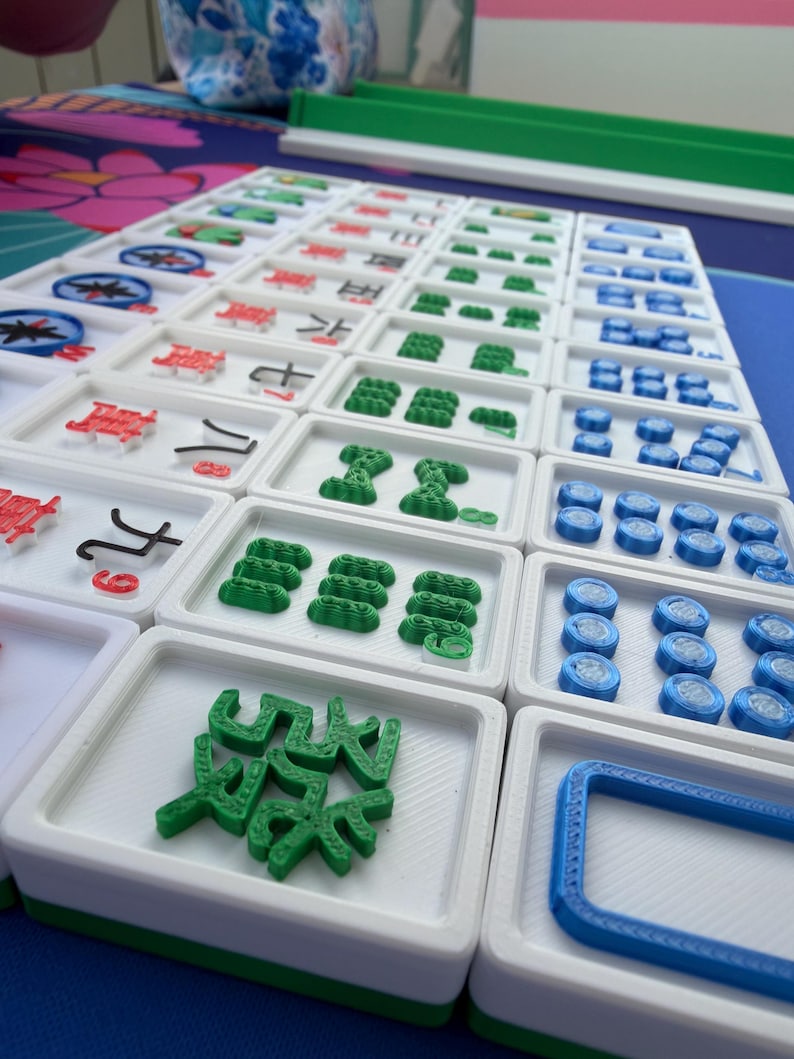 May include: A set of Mahjong tiles arranged on a blue surface. The tiles are white with green, red, and blue accents. The game pieces feature various symbols and shapes, including Chinese characters and geometric designs. The set is ready for play.