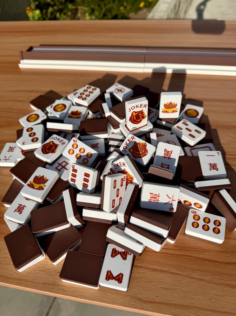May include: A collection of Mahjong tiles, featuring white faces with red and brown symbols and characters. Some tiles display the word "Joker" and various suit symbols. The tiles are stacked on a wooden surface.