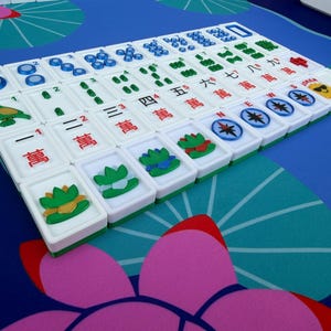May include: A close-up of a Mahjong set with white tiles featuring Chinese characters and symbols. The tiles are arranged on a blue surface with a floral pattern. The set includes green and white racks.