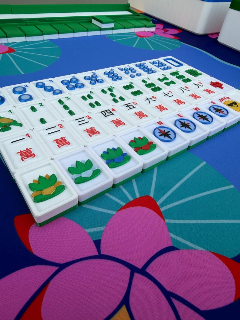May include: A close-up of a Mahjong set with white tiles featuring Chinese characters and symbols. The tiles are arranged on a blue surface with a floral pattern. The set includes green and white racks.