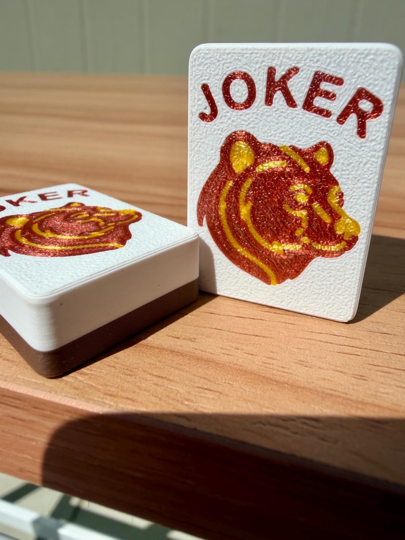 May include: White playing card with the word "JOKER" in red lettering and a red and yellow bear illustration. A deck of cards with a brown border is also visible.