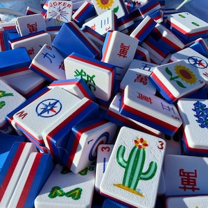 May include: A collection of white, blue, and red Mahjong tiles. The tiles feature various symbols, numbers, and illustrations, including a sunflower, cactus, and compass rose. The tiles are stacked together, creating a colorful and engaging display.