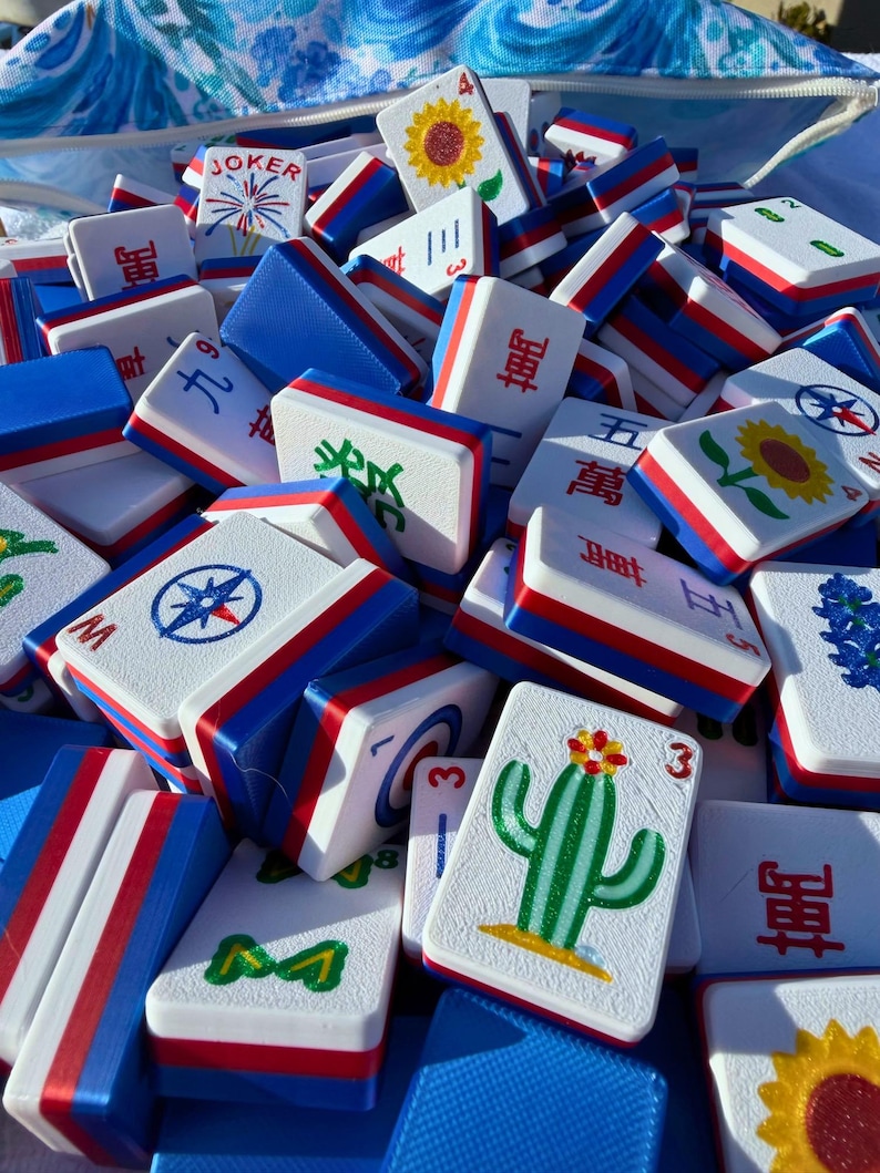 May include: A collection of white, blue, and red Mahjong tiles. The tiles feature various symbols, numbers, and illustrations, including a sunflower, cactus, and compass rose. The tiles are stacked together, creating a colorful and engaging display.