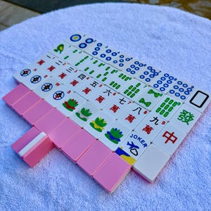 May include: A set of Mahjong tiles arranged on a white towel. The tiles are primarily white with various symbols and characters in green, blue, and red. Pink tiles are also present, with some labeled "JOKER".
