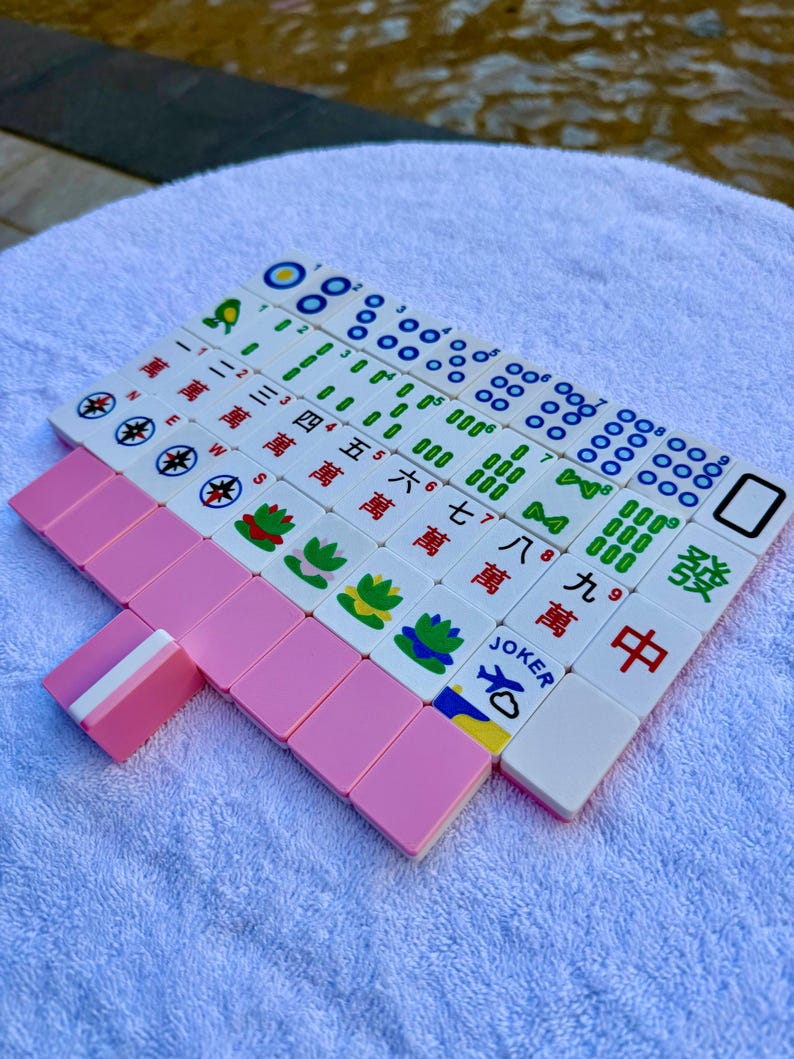 May include: A set of Mahjong tiles arranged on a white towel. The tiles are primarily white with various symbols and characters in green, blue, and red. Pink tiles are also present, with some labeled "JOKER".