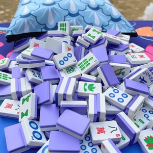 May include: A pile of white and lavender Mahjong tiles with various symbols and numbers. The tiles are scattered in front of a blue patterned pillow. The game pieces are for the traditional tile-based game.