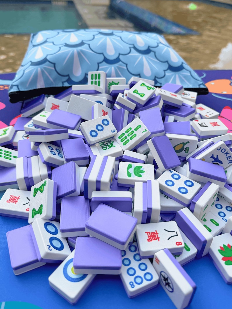 May include: A pile of white and lavender Mahjong tiles with various symbols and numbers. The tiles are scattered in front of a blue patterned pillow. The game pieces are for the traditional tile-based game.
