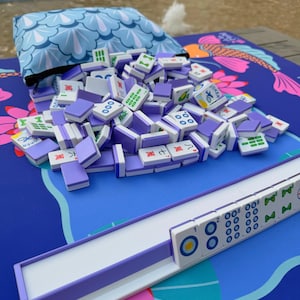 May include: A set of Mahjong tiles in purple and white, spilling from a blue pouch with a wave pattern. The tiles are scattered on a blue mat with a koi fish design. A white and purple tile rack is also visible.