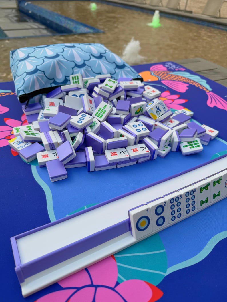 May include: A set of Mahjong tiles in purple and white, spilling from a blue pouch with a wave pattern. The tiles are scattered on a blue mat with a koi fish design. A white and purple tile rack is also visible.