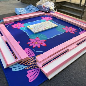 May include: A Mahjong set with pink and white tile holders and a blue mat featuring a koi fish and lotus flower design. The set includes a tile rack and a small tile holder with various symbols.