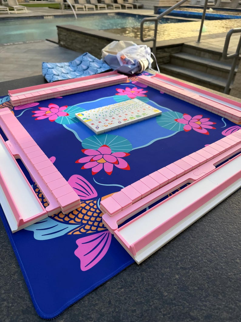 May include: A Mahjong set with pink and white tile holders and a blue mat featuring a koi fish and lotus flower design. The set includes a tile rack and a small tile holder with various symbols.