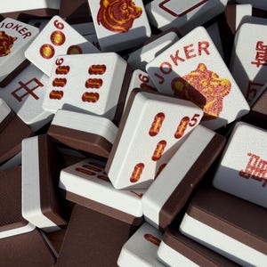 May include: A collection of white and brown Mahjong tiles. Some tiles feature red symbols, numbers, and the word "JOKER" with a bear illustration. The tiles are stacked together, showcasing the game's components.