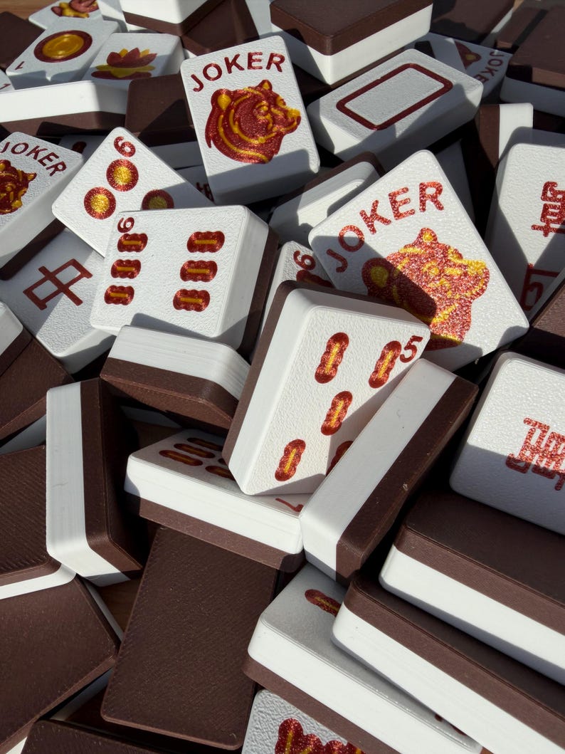May include: A collection of white and brown Mahjong tiles. Some tiles feature red symbols, numbers, and the word "JOKER" with a bear illustration. The tiles are stacked together, showcasing the game's components.