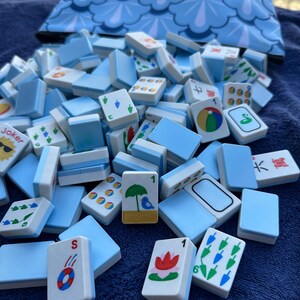 May include: A collection of light blue and white Mahjong tiles with various symbols and numbers, scattered on a blue surface. A matching blue patterned bag is in the background. The tiles feature red, green, and blue designs.