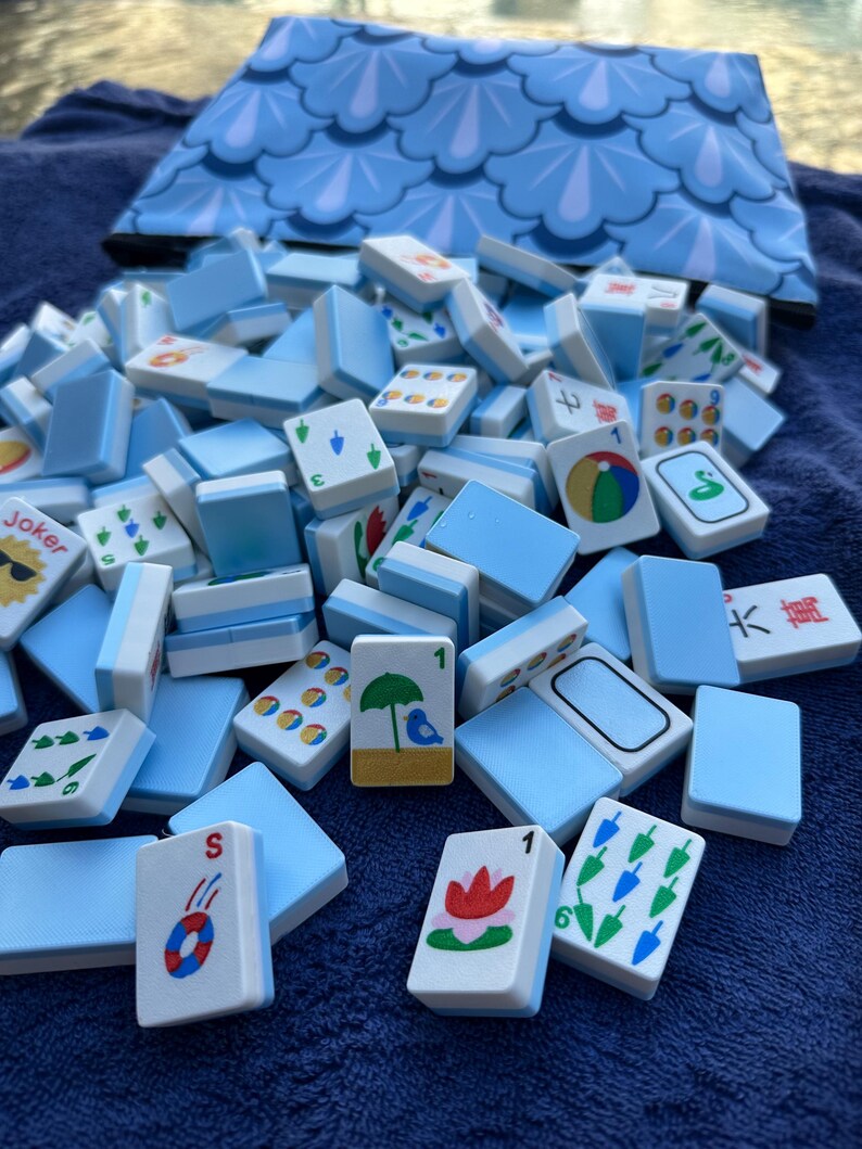 May include: A collection of light blue and white Mahjong tiles with various symbols and numbers, scattered on a blue surface. A matching blue patterned bag is in the background. The tiles feature red, green, and blue designs.