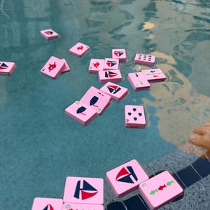 May include: Pink Mahjong tiles with various symbols, including sailboats and fish, floating in a pool. The tiles are scattered across the water, creating a playful and colorful scene. The water's surface reflects the sunlight, adding to the vibrant aesthetic.