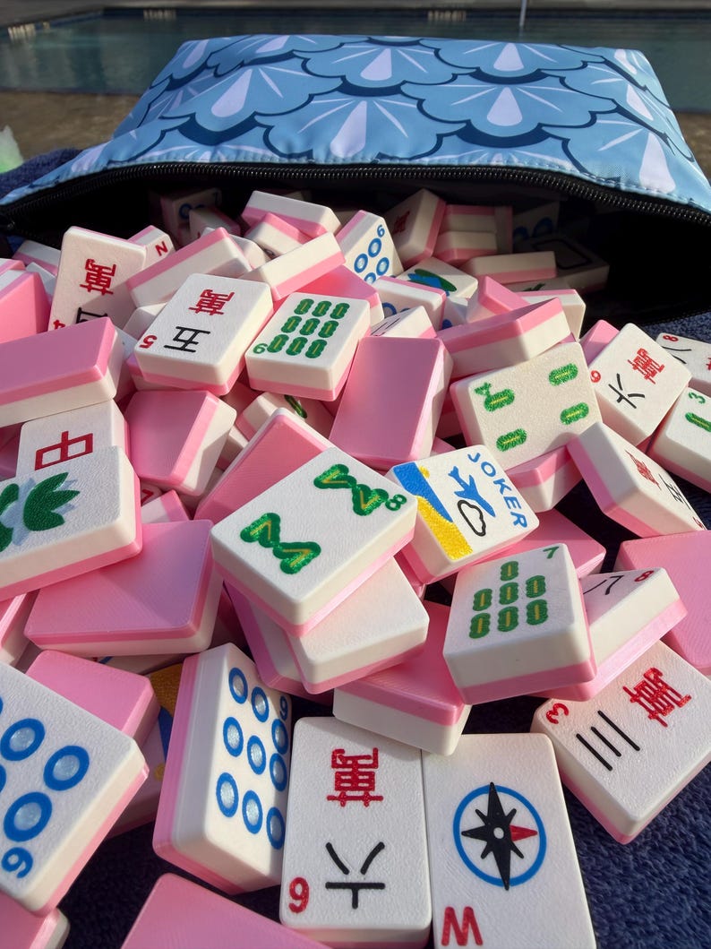 May include: A pile of pink and white Mahjong tiles spilling out of a blue zippered bag with a white and blue scale pattern. The tiles have various symbols and numbers in black, green, and blue.