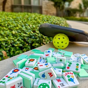 May include: A collection of white and green Mahjong tiles with various symbols and Chinese characters, scattered on a concrete surface. A yellow pickleball and a black paddle rest in the background, suggesting outdoor recreational activity.