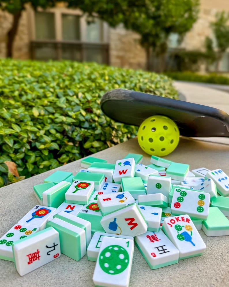 May include: A collection of white and green Mahjong tiles with various symbols and Chinese characters, scattered on a concrete surface. A yellow pickleball and a black paddle rest in the background, suggesting outdoor recreational activity.